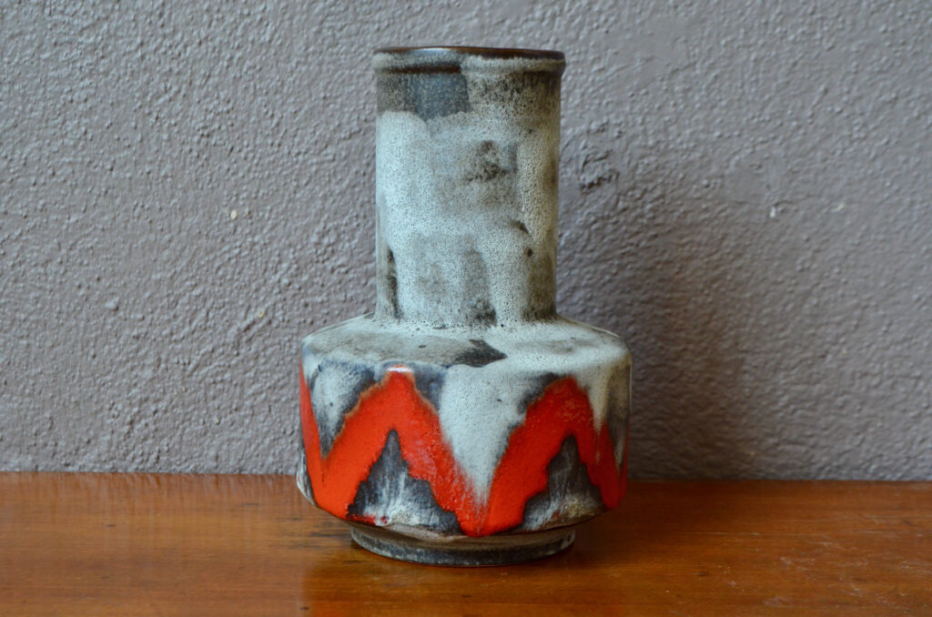 Production ouest-allemande vintage, ce vase à la forme originale est typique des productions Fat Lava. Sur un fond grisé, minéral nuancé et texturé apparaissent des zébrures rouge à l'aspect tribal.  Surprenante touche déco colorée, cette céramique saura trouver sa place sur une belle enfilade ou une commode chinée.