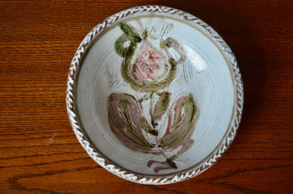 Cette belle coupe en terre chamottée est une production du potier Albert Thiry à Vallauris. C'est une pièce touchante, sophistiquée et rustique à la fois. Le motif central est souligné par un liseré verre et des scarifications sur le tour de la pièce. Le travail de la terre chamottée, typique du céramiste apporte sont grain et sa texture à la céramique. Plat de service, décoration de centre de table ou vide poche, c'est une pièce émouvante inspirée par l'art populaire.