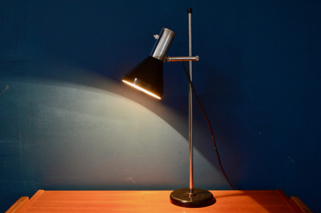 Le design de cette petite lampe est touchant de simplicité et d'élégance. Ses lignes sobres et fonctionnelles l'ancrent dans le design des années 60. Réglable en hauteur et orientable, elle est aussi pratique que sobre. Le chrome du support se marie à merveille avec la peinture sombre du déflecteur. Nous aimons l'imaginer sur une table à dessin d'architecte ou comme compagne de bricolage ou d'écriture.